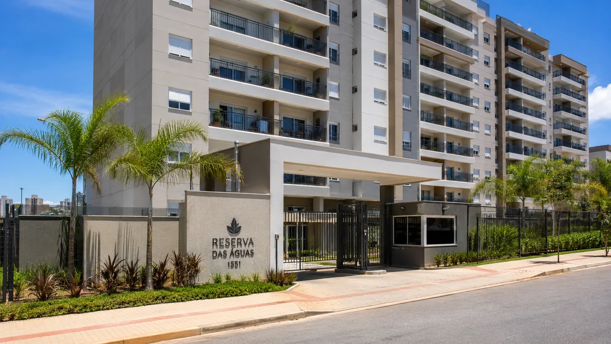 Apartamentos. Fachada de um condomínio de apartamentos residenciais moderno com sacadas de vidro e portaria, cercado por árvores em um dia ensolarado no Brasil